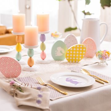 Show details for Natural Placemat (2-Pack, Tan, 12" x 18") A festive table setting with pastel Easter eggs, lit candles with colorful bases, and a white plate featuring a purple bunny design. The table is adorned with a small floral arrangement and a dish of pastel candies, creating a cheerful spring atmosphere.