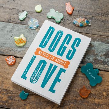 Show details for Pet Tag Mold A wooden sign with the text "DOGS NEVER LIE ABOUT LOVE" is placed on a rustic wooden surface. Surrounding the sign are colorful, pet-shaped tags including paw prints and bones, with one tag labeled "Luna.