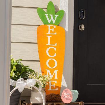 Show details for Carrot Leaner A wooden carrot-shaped sign with the word "WELCOME" leans against a wall near a black door. Next to it, there are potted plants and a small, round sign that reads "Happy Spring.
