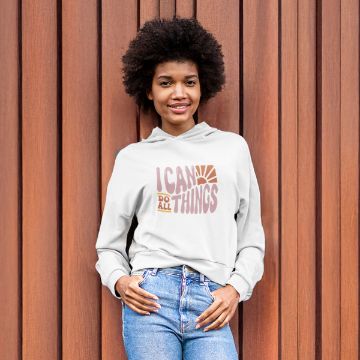 Show details for Do All Things A person with curly hair is smiling and wearing a light-colored hoodie with the text "I CAN DO ALL THINGS" printed on the front. They are standing in front of a wooden wall.