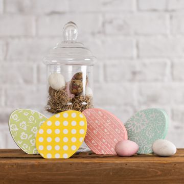 Show details for Easter Egg Patterns Decorative Easter eggs on a wooden table. Three large eggs with patterns—yellow with dots, pink with stripes, and green with floral design—are in front. A glass jar filled with moss and eggs is in the background against a white brick wall.