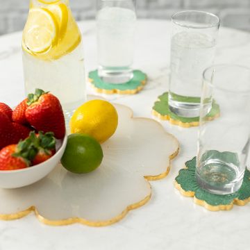 Show details for Tropical Tray & Coasters Molds Bundle A table with a decorative placemat holds a jug of lemon water and three glasses. A bowl of strawberries, a lemon, and a lime rest alongside. Green coasters with gold edges accompany the glasses on the white marble surface.