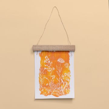 Show details for Watercolor Paper (600 gsm, 8½" x 11") A piece of art featuring white floral and plant designs on an orange and yellow gradient background. It's hung by a twisted string and a wooden clip against a beige wall.
