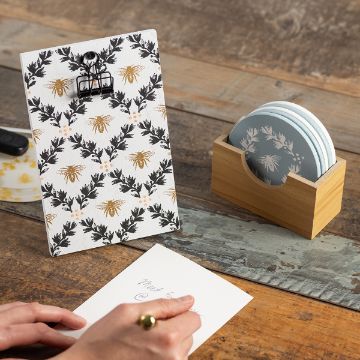 Show details for Clipboard Stand A person is writing on a paper with a pen at a wooden table. A clipboard with a floral and bee pattern and a wooden holder with coasters beside it are on the table.