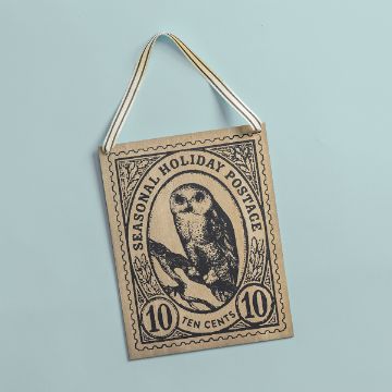 Show details for Hanging Sign (8½" x 11") A decorative plaque shaped like a postage stamp features an illustration of an owl with the text "Seasonal Holiday Postage" and "Ten Cents." It has a ribbon for hanging and is set against a light blue background.