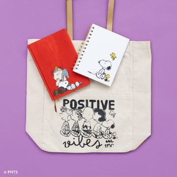 Show details for Peanuts - Cuddle Puddle A beige tote bag with "Positive Vibes" and cartoon characters is laid on a purple background. On top of the bag are two notebooks: a red one with a character resting on it and a white spiral notebook with characters on its cover.