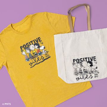 Show details for Peanuts - Positive Vibes Yellow t-shirt and beige tote bag on a purple background, both featuring cartoon characters and the text "Positive Vibes" with the year "1950" below.