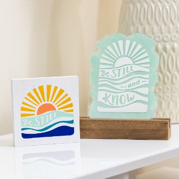 Show details for Be Still And Know Two decorative signs on a shelf: one square with a colorful sun, waves, and "Be Still"; the other with a sun and "Be Still and Know" on green acrylic, set in a wooden stand. Cream-colored, textured background.
