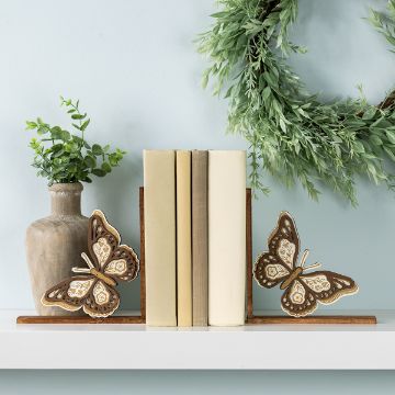 Show details for Floral Butterfly A shelf displays books held upright by decorative butterfly bookends. To the left is a rustic vase with green foliage. Above, a leafy wreath hangs on the light blue wall.
