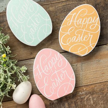 Show details for Happy Easter Egg Decorative Easter eggs with "Happy Easter" written in elegant script on pastel backgrounds (green, orange, pink) sit on a wooden surface. Nearby, two plain eggs and small sprigs of greenery with yellow flowers add to the festive scene.