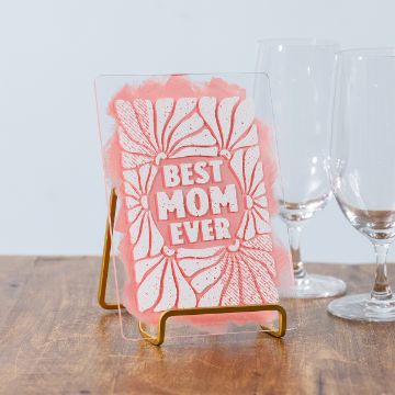 Show details for Best Mom Ever A card with "Best Mom Ever" text in bold white letters surrounded by pink and white floral patterns sits on a wooden surface. It's supported by a gold stand, with three empty wine glasses in the background.