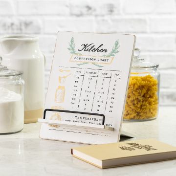 Show details for Kitchen Conversion Chart A kitchen conversion chart displaying measurements for cups, fluid ounces, tablespoons, and teaspoons, along with temperature conversions. It stands on a white countertop beside jars of sugar and pasta, with a cookbook nearby.
