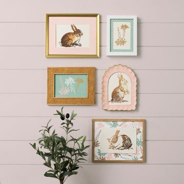 Show details for Bunny Trio A collection of five framed art pieces featuring rabbits and flowers arranged on a light-colored wall. The frames vary in shape and color, with some in gold and others in pastel shades. A plant is visible at the lower left corner.