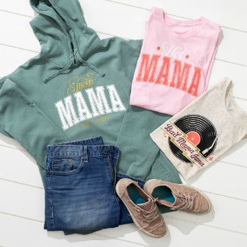 Show details for Bad Mama Jama Flat lay of casual clothing items on a white surface: a green hoodie with "Tired Mama" text, a pink shirt with "Hot Mama" text, a beige T-shirt with a record design, blue jeans, and brown sneakers.