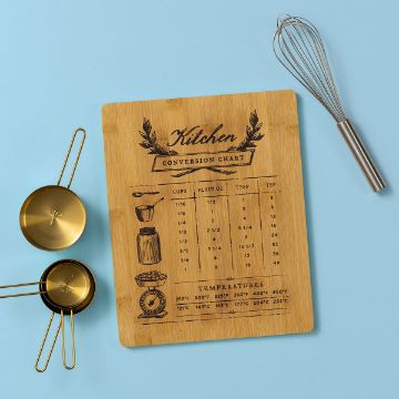 Show details for Bamboo Board (9" x 11¾") A bamboo cutting board with a kitchen conversion chart engraved on it is placed on a blue surface. Nearby are two brass measuring cups and a metal whisk. The chart includes measurements for cups, fluid ounces, tablespoons, and temperatures.