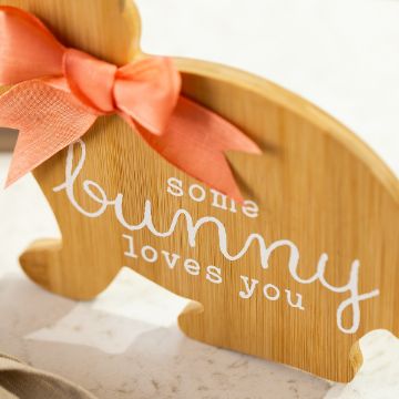 Show details for Somebunny Loves You Wooden bunny-shaped decoration with a coral bow around its neck. The text on the bunny reads "some bunny loves you" in white letters. The background is a light-colored surface.