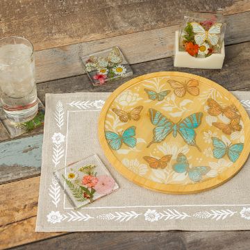 Show details for Square Coaster Set Bundle A round wooden board with colorful butterfly designs is on a linen placemat with floral embroidery. Glass coasters with pressed flowers are scattered around, alongside a glass of water with ice. A small box holds more flower-adorned coasters.