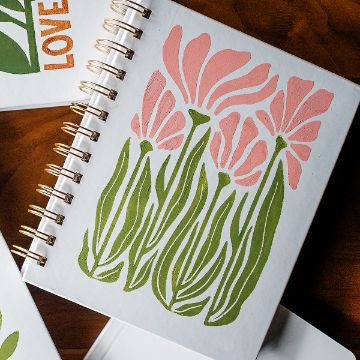 Show details for Wild Flowers A spiral-bound notebook with a cover illustration of pink flowers and green leaves, set on a wooden surface. Nearby, parts of other notebooks with similar designs and the word "LOVE" partially visible.