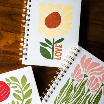 Show details for Love Blooms Three spiral-bound notebooks with floral illustrations on their covers are arranged on a wooden table. One features a yellow flower with the word "LOVE," another has pink flowers, and the third has a red and green design.