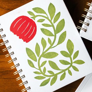 Show details for Wandering Vine A spiral-bound notebook with a white cover featuring a stylized illustration of a red flower and green leafy stem. The notebook is placed on a wooden surface beside another partially visible notebook.