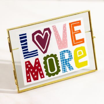 Show details for Love More A colorful framed artwork with the phrase "LOVE MORE" featuring letters in various patterns and colors, including stripes, hearts, polka dots, and solid colors. The frame is gold and transparent, placed on a light surface.