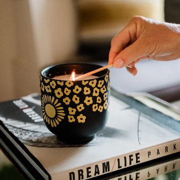 Show details for Full Bloomin’ A hand lighting a candle in a black holder decorated with yellow flowers. The candle is placed on two stacked books, with one titled "DREAM/LIFE PARK." The scene is cozy and warm.