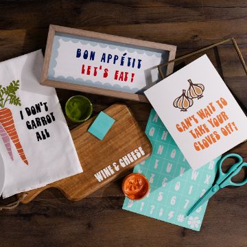 Show details for Tiff’s Font A top view of a wooden table with items including a "Bon Appétit, Let's Eat!" framed sign, a "Can't wait to take your cloves off!" garlic card, a "I don't carrot all" tea towel, a wooden board labeled "Wine & Cheese," scissors, and alphabet stickers.