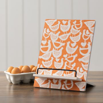 Show details for Hen House A patterned orange cookbook stand with white illustrations of chickens. It rests on a wooden table next to a carton holding nine brown eggs. The background is a white paneled wall.