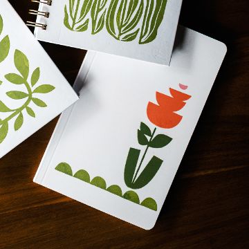 Show details for Funky Shapes Three notebooks with botanical designs on a dark wooden surface. One notebook features abstract green leaves, another has a leafy pattern, and the third displays a stylized red flower with green leaves and a pattern of green dots at the bottom.