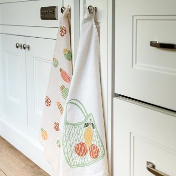 Show details for Tote-ally Two white kitchen towels hang on silver knobs. One towel features colorful fruit designs, and the other shows a green net bag with orange and yellow fruit. The backdrop is a white paneled kitchen cabinet with a light wood floor.