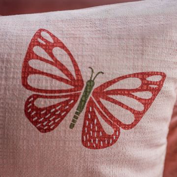 Show details for Butter-flyin’ A close-up image of a cushion with a red butterfly design on a light pink fabric. The butterfly has a green body with intricately patterned wings, featuring white dotted accents. The background is softly textured.