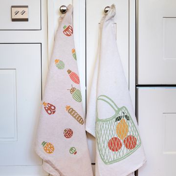 Show details for Buggin’ Out Two white kitchen towels hang on knobs against a white cabinet. The left towel features colorful kitchen utensil designs, while the right towel has a green mesh bag pattern with red and orange fruit inside.