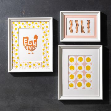 Show details for Ham And Eggs Three framed artworks on a wall: one with the phrase "EGG DEALER" and a chicken silhouette, one with patterned yellow circles resembling eggs, and one with stylized strips of bacon on a pink background.