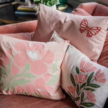 Show details for Bunch Of Blossoms Three decorative pillows on a pink chair. One pillow features a red butterfly, another displays stylized pink flowers with green leaves, and the third has large pink floral designs with a central white shape. A table with books is in the background.