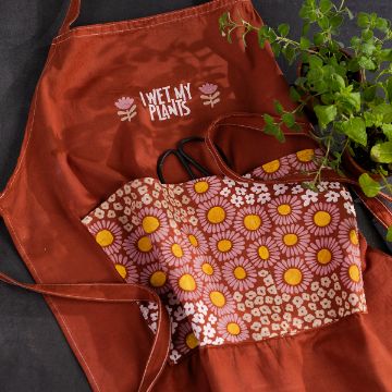 Show details for Cotton Apron (White) A terracotta-colored apron with the words "I Wet My Plants" and flower designs, paired with gardening gloves featuring a floral pattern of daisies and sunflowers. A small green potted plant is placed next to them on a dark surface.