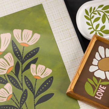 Show details for Weavin’ A flat lay image showcasing a green floral print, a black and white tray with a leaf design, and a wooden-framed art piece featuring a white flower and the word "LOVE" in pink on a brown background.