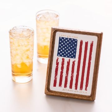 Show details for Distressed Flag Two iced drinks next to a framed print featuring a stylized American flag with text "Land of the Free" and "Because of the Brave" at the bottom. The flag has a rustic, painted appearance.