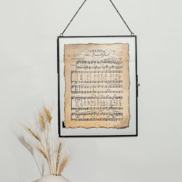 Show details for America The Beautiful A framed sheet of music titled "America the Beautiful" hangs against a light wall. The sheet has an aged appearance. A decorative vase with dried grasses is placed in the bottom left corner.