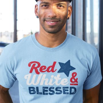 Show details for & Blessed A person wearing a light blue t-shirt with the text "Red White & Blessed" in red, white, and blue. They are standing by a window, smiling, with short hair and a cap. The background is slightly blurred.