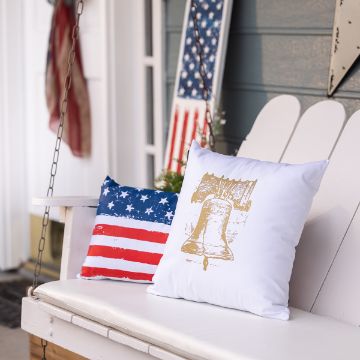 Show details for Liberty Bell Patriotic-themed porch with two pillows on a white swing. One pillow features the American flag design, and the other depicts the Liberty Bell. An American flag leans against the blue siding wall behind the swing.