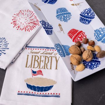 Show details for Sweet Land of Liberty Festive kitchen towels and a tray with patriotic designs, including fireworks, cupcakes, and an American flag. A towel reads "Sweet Land of Liberty." Nuts are placed on the tray. The color scheme features red, white, and blue.