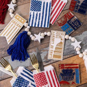 Show details for American Made Minis A festive arrangement featuring red, white, and blue-themed napkin holders with American flag designs. Surrounding décor includes beaded garland with stars, patriotic cards, yarn tassels, and silverware on a rustic wooden surface.
