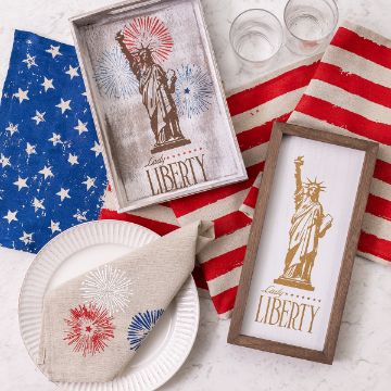 Show details for Lady Liberty Decor with Statue of Liberty images and "Lady Liberty" text on framed prints. The setup includes an American flag napkin, blue star-patterned napkin, white plate, glass cups, and a beige napkin with fireworks design, all on a light surface.