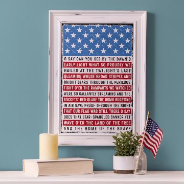 Show details for National Anthem Framed artwork on a shelf features a flag-themed background with text from the U.S. National Anthem. A small American flag is in a vase next to the frame, and candles are placed nearby. The background wall is a soft blue-green color.