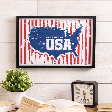 Show details for Made in the USA A framed poster with a blue map of the USA on a red and white striped background reads "Made in the USA." Below, a small potted plant and a vintage clock sit on a white shelf, alongside an open book.