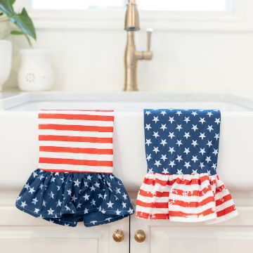 Show details for Patriotic Patterns Two kitchen towels hang over a white sink. One towel has red and white stripes, and the other has white stars on a blue background. Each towel has a ruffled bottom edge. A brass faucet and potted plant are visible in the background.