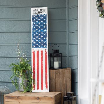 Show details for Patriotic Home A vertical wooden sign leans against a blue wall, featuring a painted American flag and the text "Welcome to our Home." Beside it, a potted plant and a lantern sit on a wooden crate, creating a cozy porch scene.