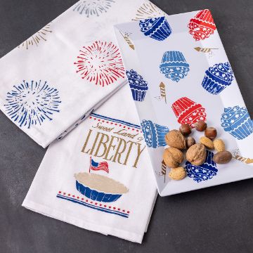 Show details for Melamine Platter (13⅝" x 7⅜") Patriotic-themed kitchen items on a dark surface, featuring a towel with fireworks and "Sweet Land of Liberty" text, alongside a rectangular tray with cupcake and firework designs in red and blue. A handful of nuts is placed on the tray.