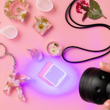 Show details for UV Flashlight Person holding a UV light over resin pendants and keychains with dried flowers and leaves on a pink background. Various molds and finished pieces are arranged around the light.