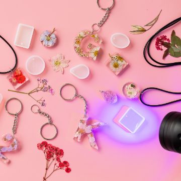 Show details for Pendant Molds Various resin keychains with dried flowers, letter shapes, and silicone molds are arranged on a pink background. A black UV flashlight shines on some molds. Loose dried flowers are scattered around.